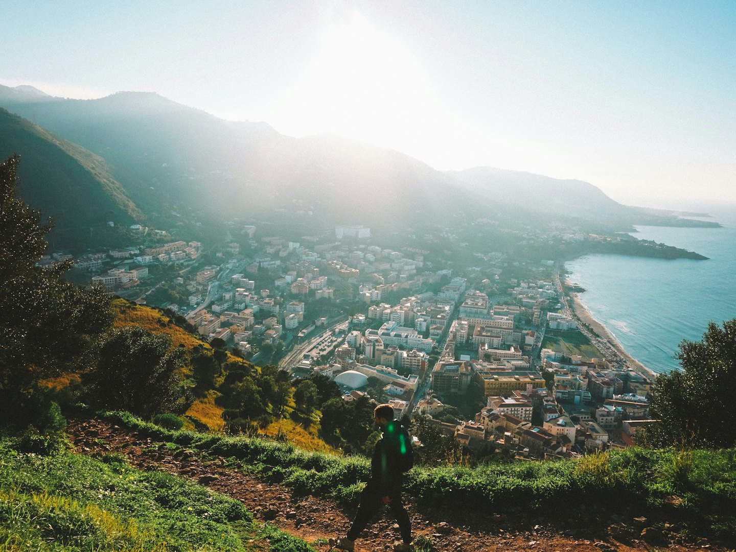 A hiker walks along a coastal hillside trail overlooking a town.