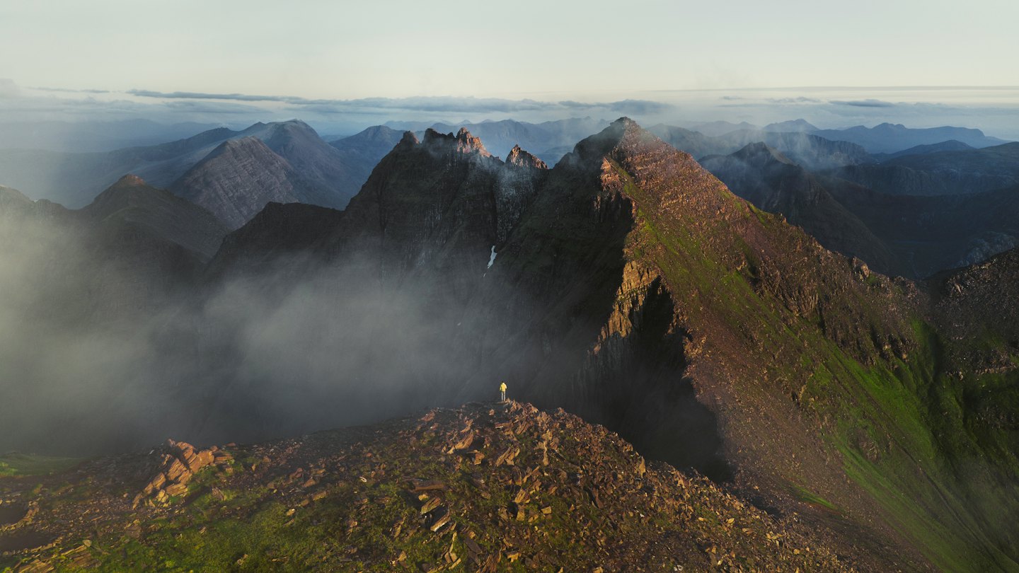 Person on a rocky mountain ridge overlooking misty peaks at sunrise.